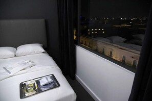 A hotel room with a bed, a tray with a bottle and glasses, and a view of buildings through the window. - Palacio San Leonardo (Puebla)