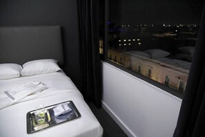 A hotel room with a bed, a tray with a glass of water and a bottle, and a window with a view of buildings at night. - Palacio San Leonardo (Puebla)