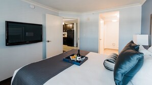 A hotel room with a bed, a flat-screen TV, a tray with snacks and a bottle, and a view of the kitchen and bathroom. - Manhattan Beach Hotel (Manhattan Beach)