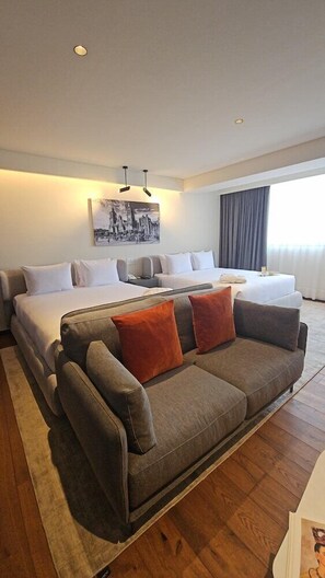 Et hotelværelse med en stor sofa, to senge, et billede på væggen og et vindue med gardiner.
