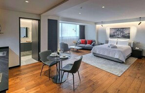 Una habitación de hotel moderna con una cama, una zona de comedor con mesa y sillas, un sofá y un ventanal grande.