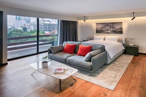 Et moderne hotelværelse med en stor seng, en grå sofa med røde puder, et lille bord og udsigt over byen gennem store vinduer.