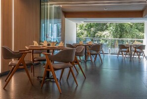 Un'area pranzo moderna con tavoli e sedie in legno, ampie finestre con vista sugli alberi e una parete divisoria in vetro.