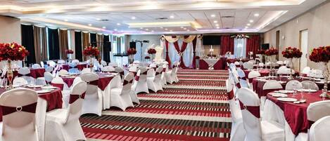Une salle de banquet avec des rangées de tables, des chaises blanches et des rideaux rouges.