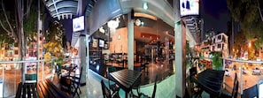Un bĂątiment moderne dotĂ© dâune façade en verre, dâun espace extĂ©rieur amĂ©nagĂ© pour les clients et dâun restaurant visible de lâextĂ©rieur.