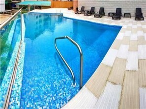 Una piscina con una escalera metálica, sillas de descanso y una zona de sol con baldosas.