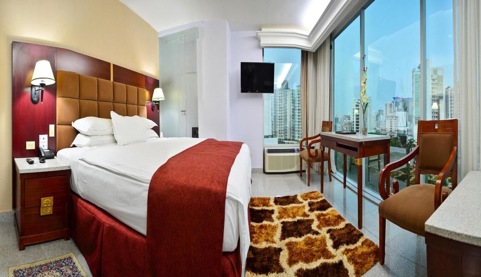 A hotel room with a bed, bedside tables, a desk, a chair, a television, and a view of a cityscape.