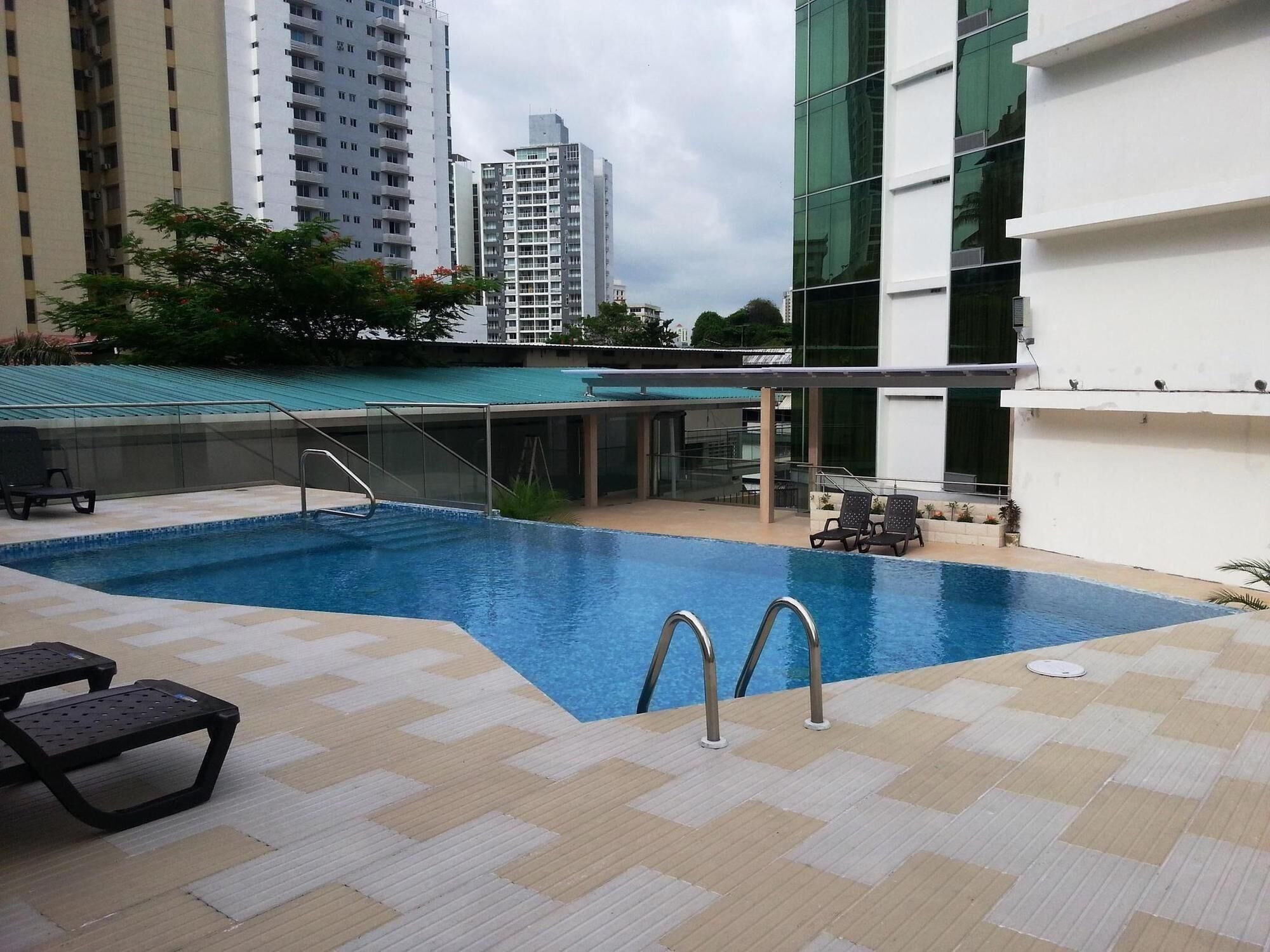A swimming pool with a tiled deck, a lounge area with chairs, and a view of surrounding buildings.