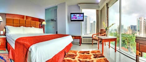A hotel room with a large bed, a television, a desk with a chair, and a view of the cityscape.