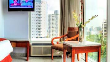 A hotel room with a bed, a TV, a chair, a table, a vase with flowers, and a view of buildings.