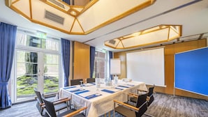 Un salón de conferencias con una larga mesa, sillas, una pantalla de proyector y vistas al exterior con vegetación.