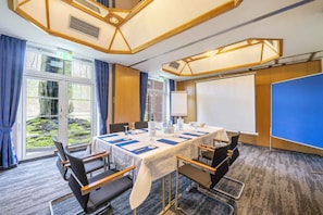 A conference room with a long table, chairs, a projector screen, and a view of greenery outside. - Hotel Schmoeker-hof (Norderstedt)