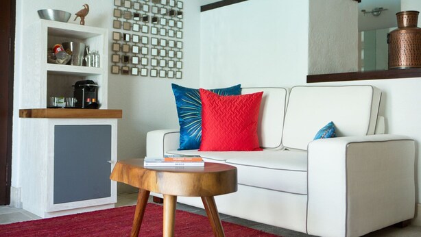 A living room with a white sofa, red and blue pillows, a wooden coffee table, a kitchen area with shelves, and a decorative wall.