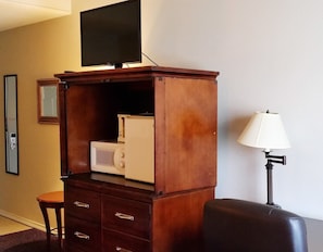 Quarto compacto com televisão sobre um armário de madeira, micro-ondas, luminária e uma mesa pequena.