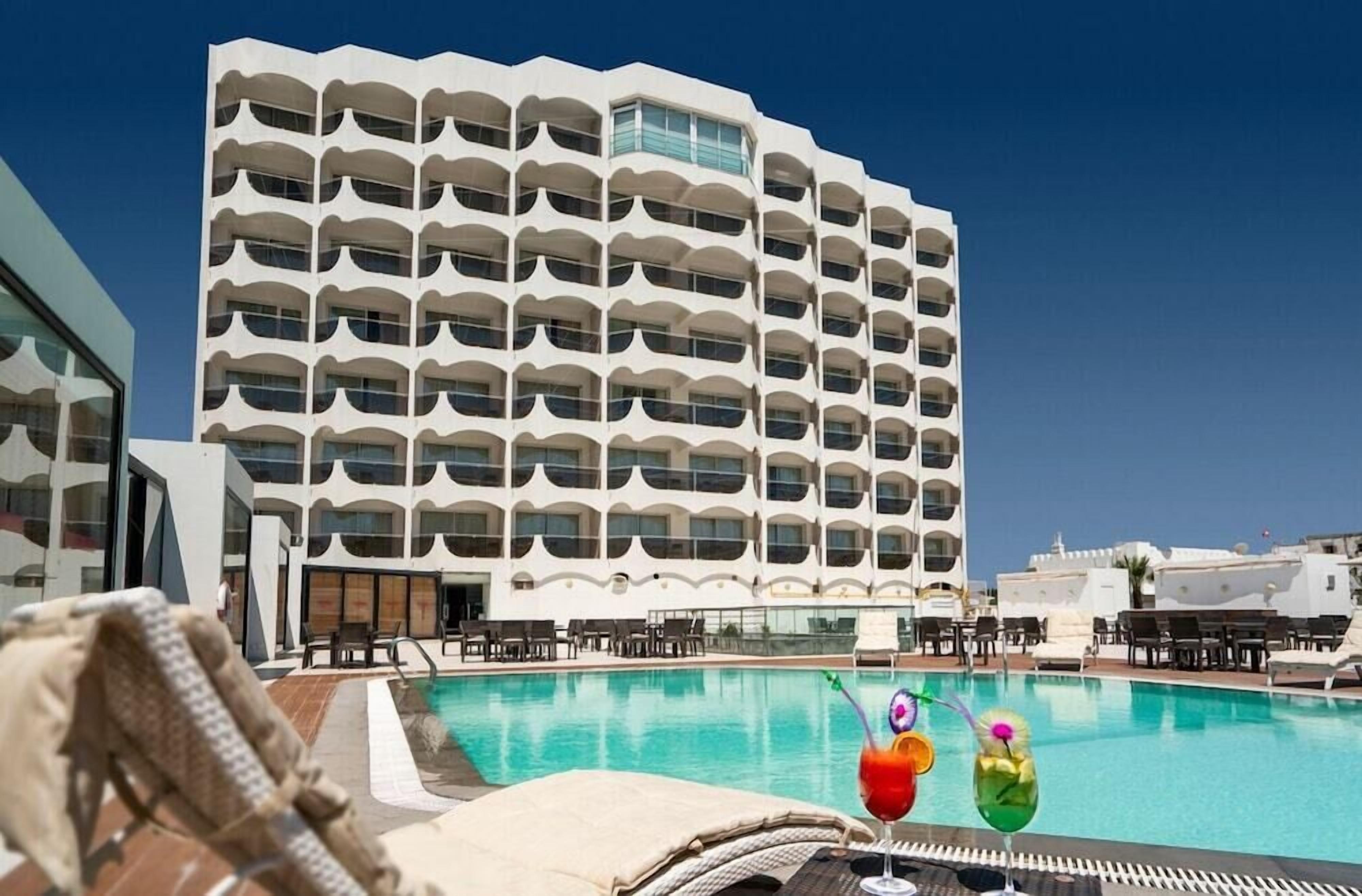 A hotel with a pool, lounge chairs, and a clear blue sky.