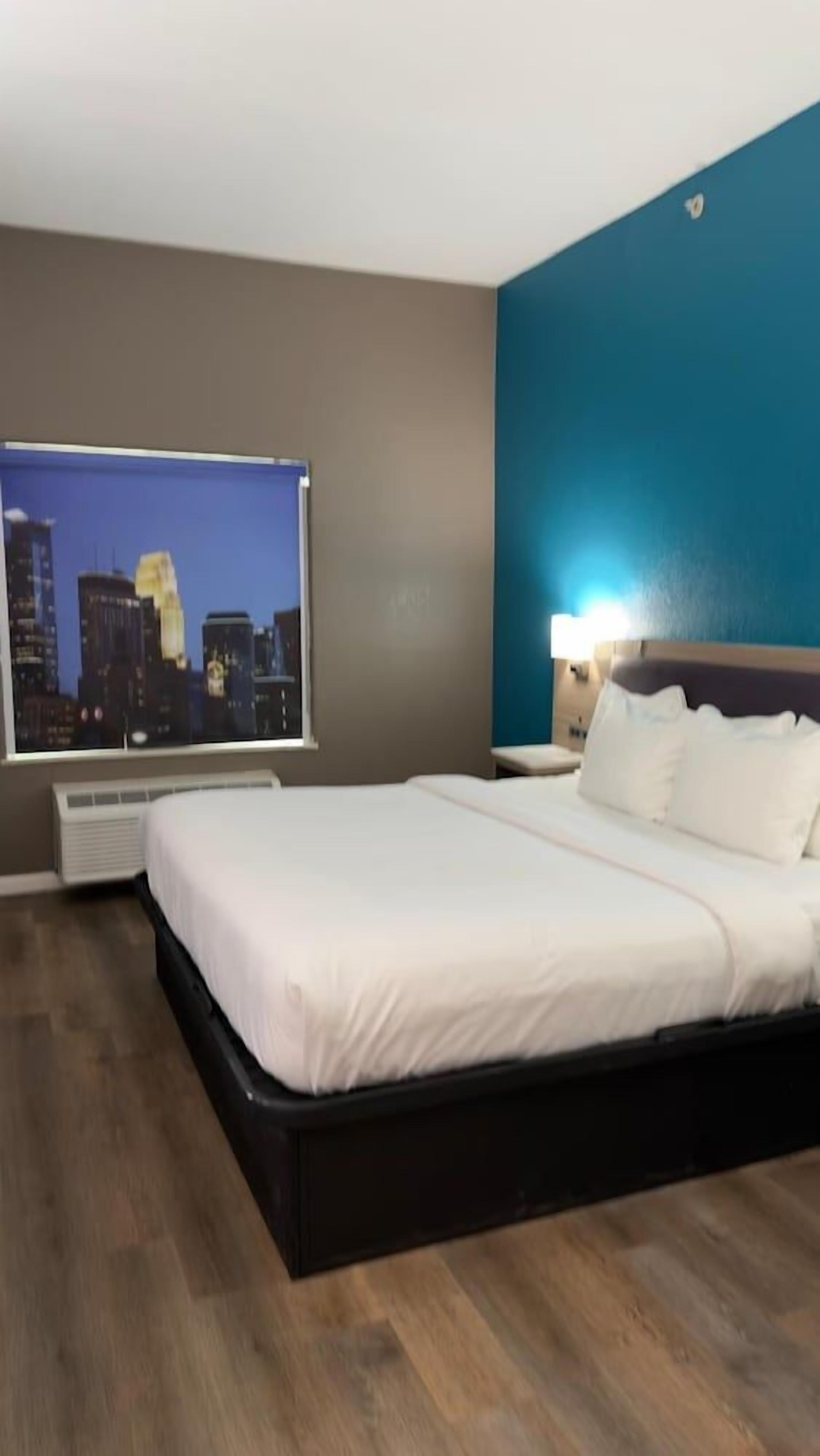 A hotel room with a bed, a cityscape view, a nightstand, and a wall-mounted air conditioning unit.