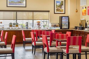 A restaurant with red upholstered chairs and tables, a coffee station with various coffee-making equipment, and framed pictures on the walls. - Red Roof Plus+ San Antonio Northeast (San Antonio)