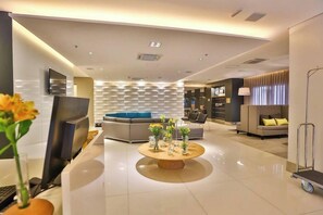 A modern hotel lobby with a round wooden coffee table, sofas, a TV, and a wall with a geometric pattern. - Roomo Santos Residencial (Santos)