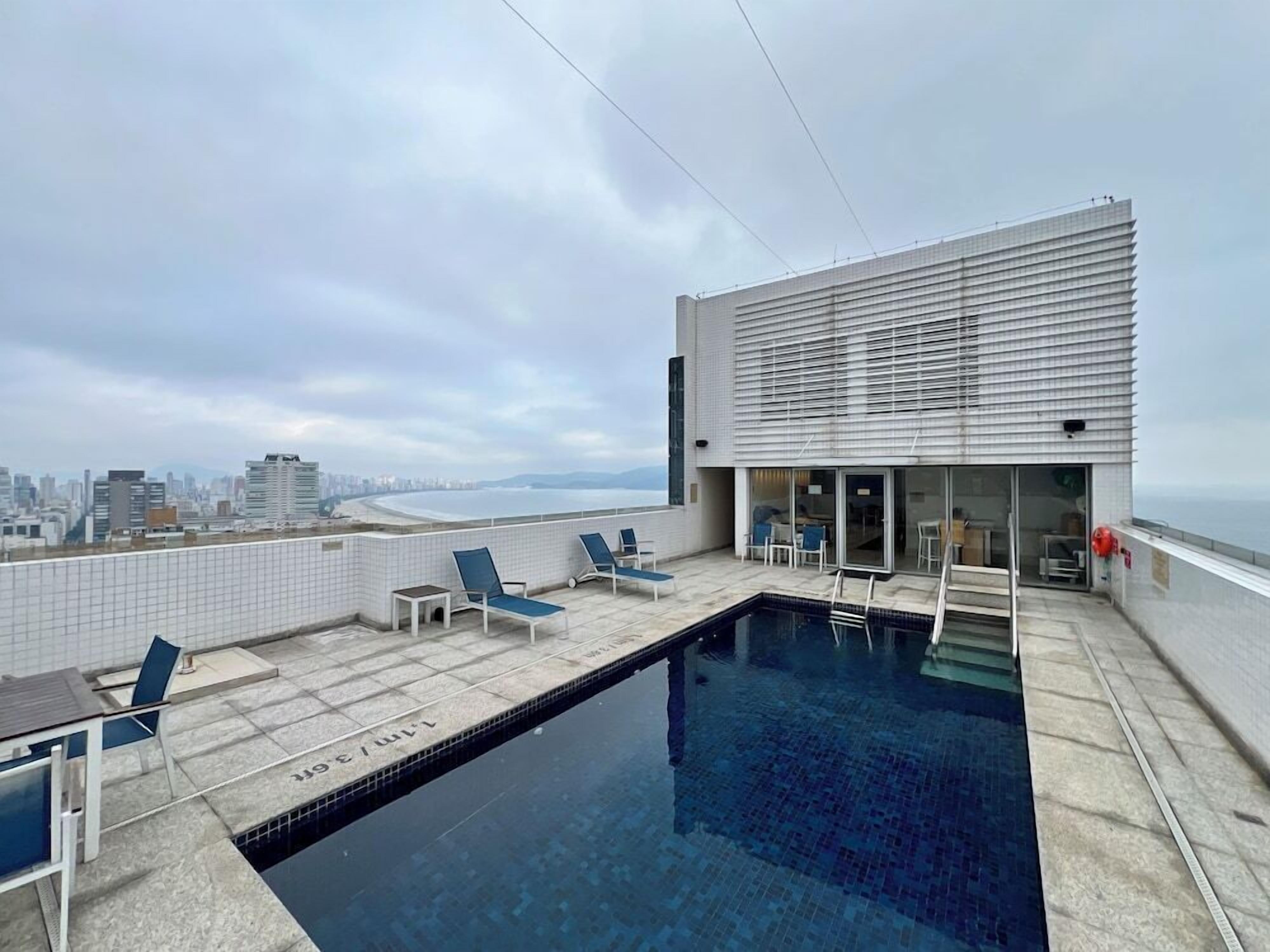 Piscina no terraço com espreguiçadeiras, vista da cidade e um edifício moderno com grandes janelas.