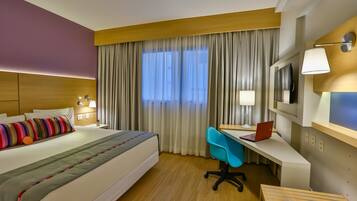 Quarto de hotel com cama, escrivaninha, cadeira e televisão.