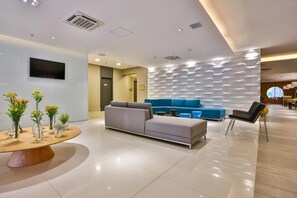 A modern lobby with a grey sofa, blue seating, a round table with vases, and a wall-mounted TV. - Roomo Santos Residencial (Santos)