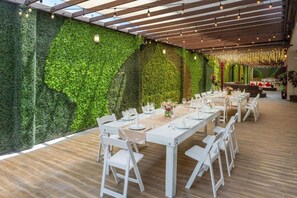 Uma sala de jantar com uma mesa comprida posta para refeição, rodeada por cadeiras brancas, parede verde e teto de madeira.