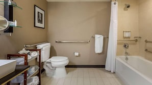 A bathroom with a toilet, a bathtub, a shower curtain, a handrail, and a shelf with towels. - Cambria Hotel Ft Collins (Fort Collins)