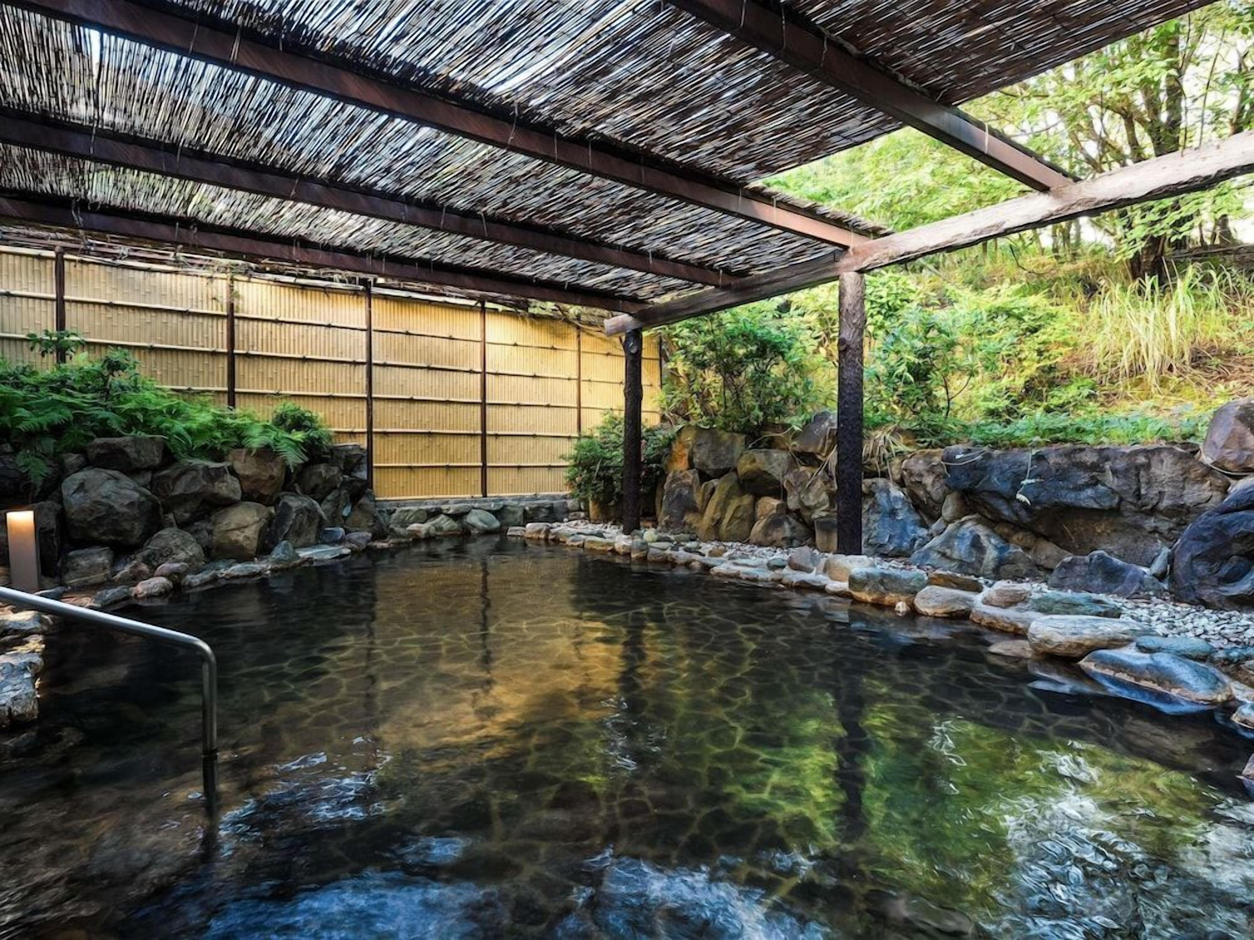 Un onsen japonés tradicional con estructura de madera, piscina revestida de piedra y pared de bambú.