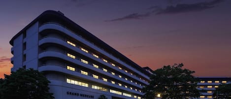 Un hotel de varios pisos con fachada curva, llamado GRAND MERCURE, que cuenta con un estacionamiento y varios árboles.
