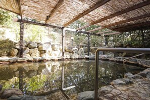 Un área tranquila de aguas termales al aire libre con una piscina de piedra, estructuras de madera y techo de paja.
