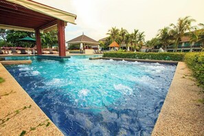 Uma banheira de hidromassagem com deck de madeira e espreguiçadeiras, rodeada por palmeiras e uma piscina.