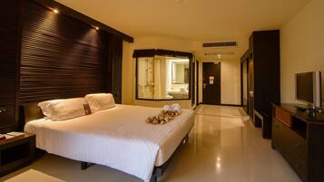 A modern hotel room with a large bed, a TV, a dresser, and a bathroom visible through a glass door.