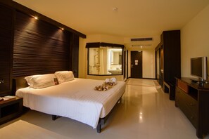 A modern hotel room with a large bed, a TV, a dresser, and a bathroom visible through a glass door.
