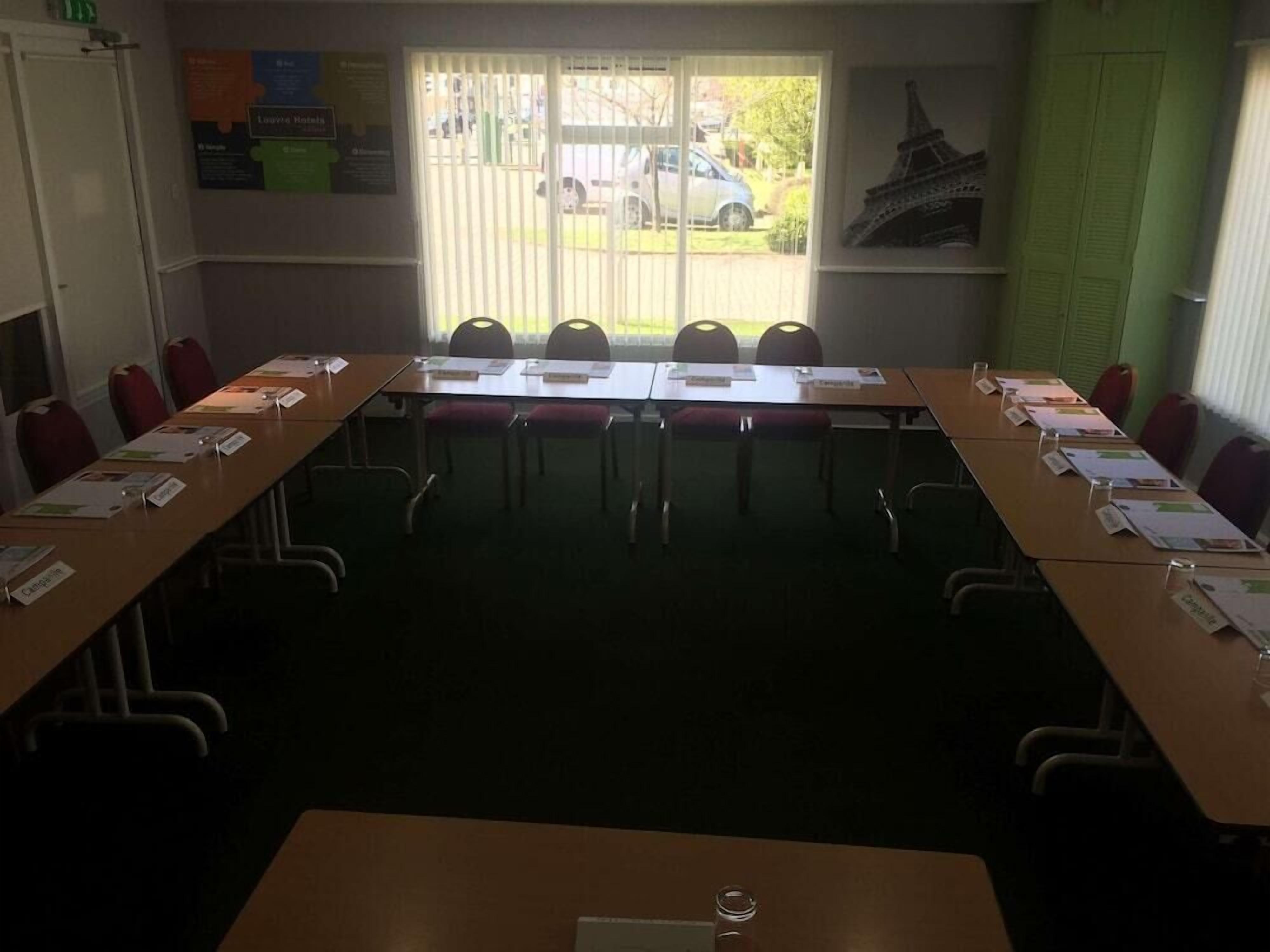 A conference room with a long table, chairs, nameplates, and a window with blinds.