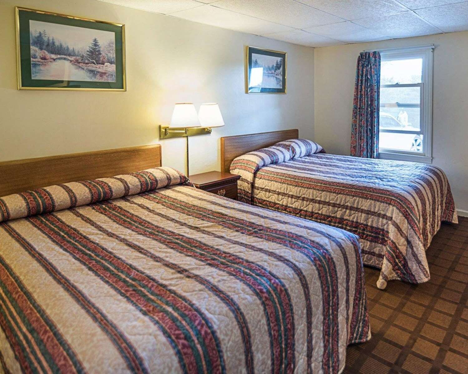 A hotel room with two beds, a nightstand, a lamp, and a window with curtains.