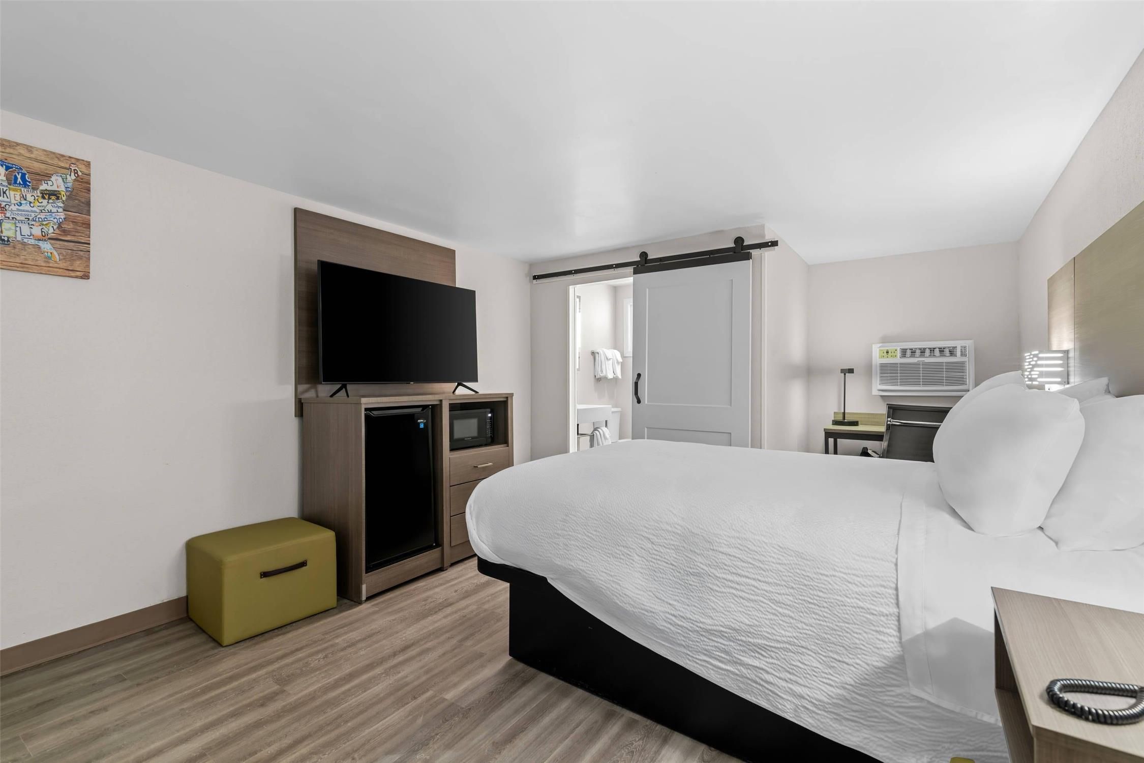 A hotel room with a bed, a television, a microwave, a small refrigerator, a desk with a lamp, and a bathroom visible through an open door.