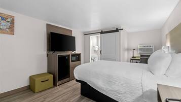 A hotel room with a bed, a television, a microwave, a small refrigerator, a desk with a lamp, and a bathroom visible through an open door.