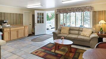 A living room with a sofa, coffee table, and a rug. There is a door leading outside, a window with a view, and a kitchen area with cabinets and a counter.
