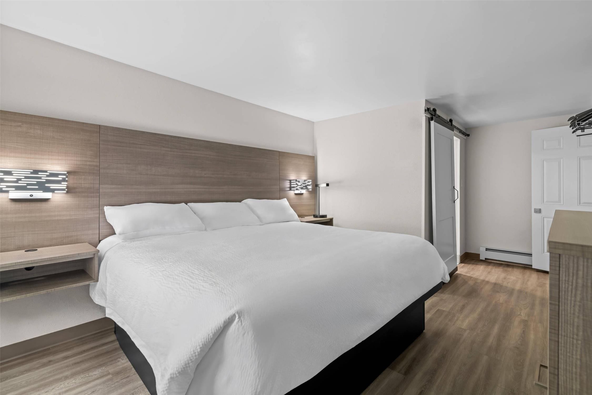 A modern bedroom with a large bed, a bedside table, a wall-mounted light, and a door.