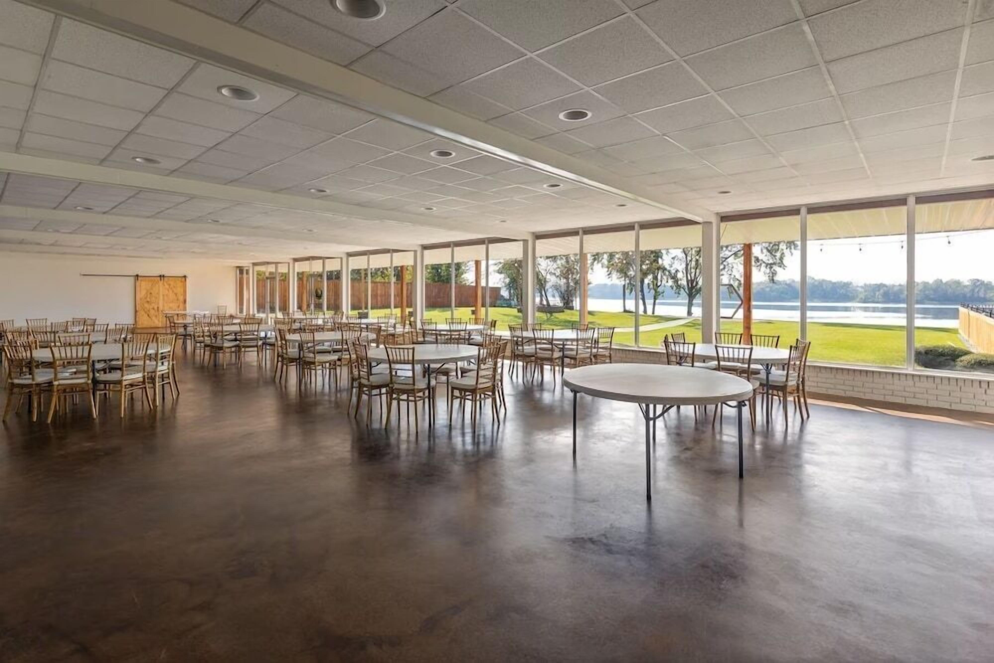 A spacious indoor area with tables and chairs arranged for dining, large windows offering a view of a lake, and a high ceiling with recessed lighting.