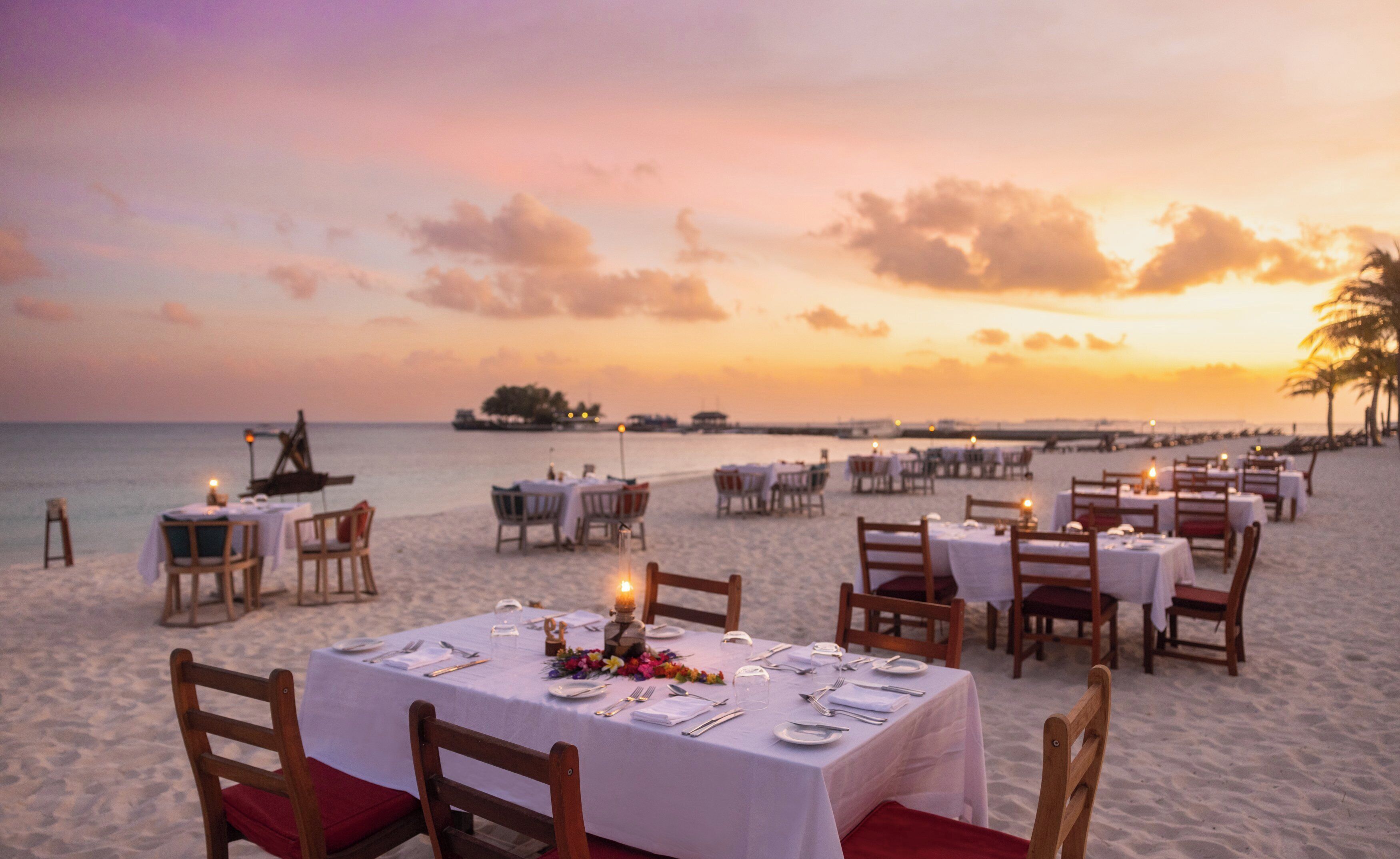 A beachside dining area with tables set for two, overlooking the ocean at sunset.