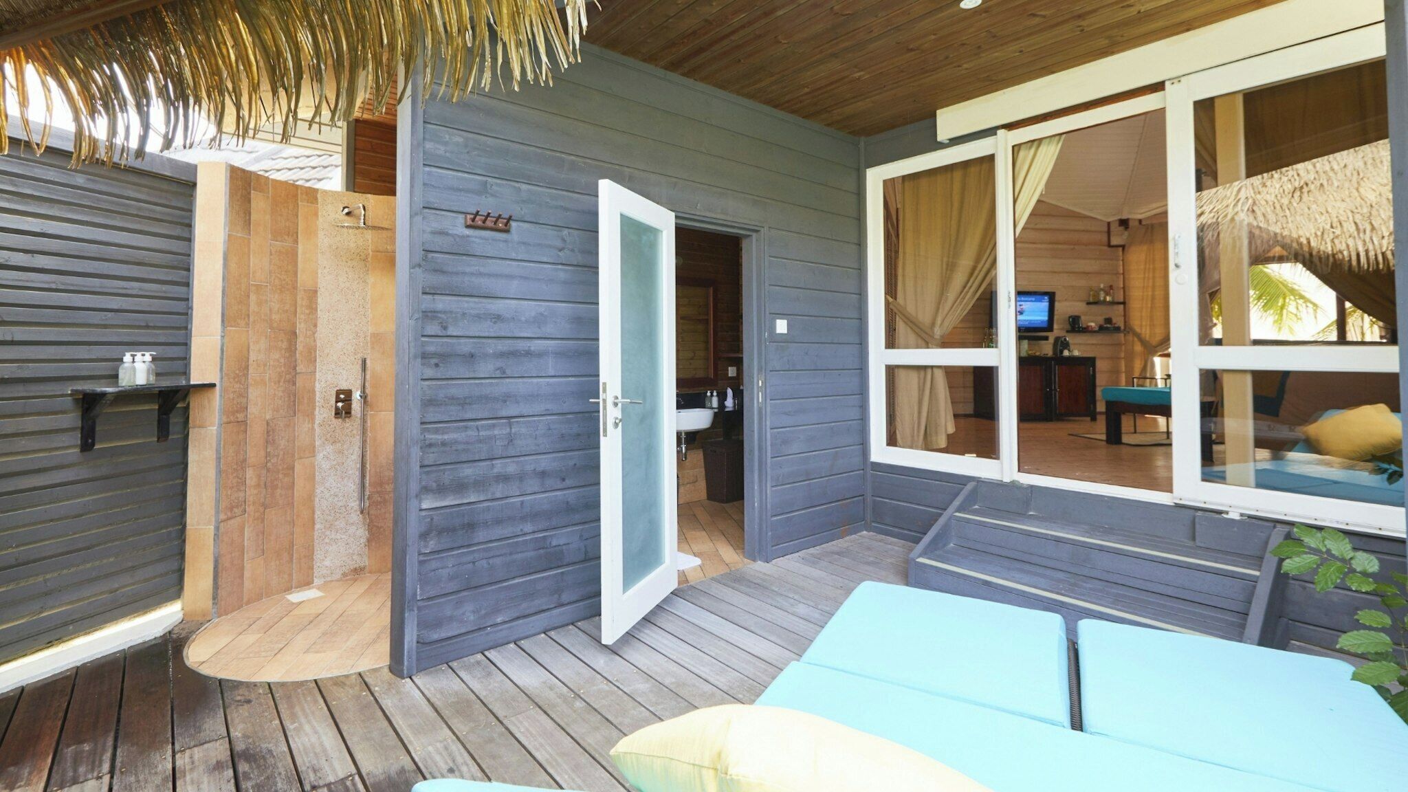 A wooden deck with a blue sofa, a door leading to a bathroom, and a TV in the adjacent room.