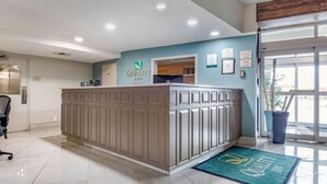 A reception desk with a sign for 'Quality Inn.' There are framed certificates on the wall, a potted plant, and a blue doormat with the hotel name. - Quality Inn Sarnia (Point Edward)