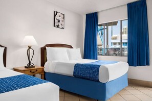 Hotellihuone, jossa on kaksi sänkyä, sinisillä verhoilla varustettu ikkuna, yöpöytä lampun kanssa ja seinälle ripustettu taulu.