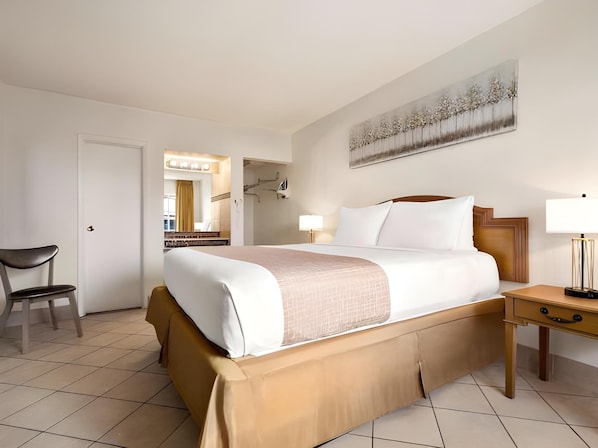 A hotel room with a bed, a chair, a nightstand, a lamp, a painting on the wall, and a door leading to a bathroom.
