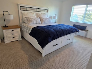 A bedroom with a white bed, a bedside table, a lamp, a window with blinds, and a carpeted floor.