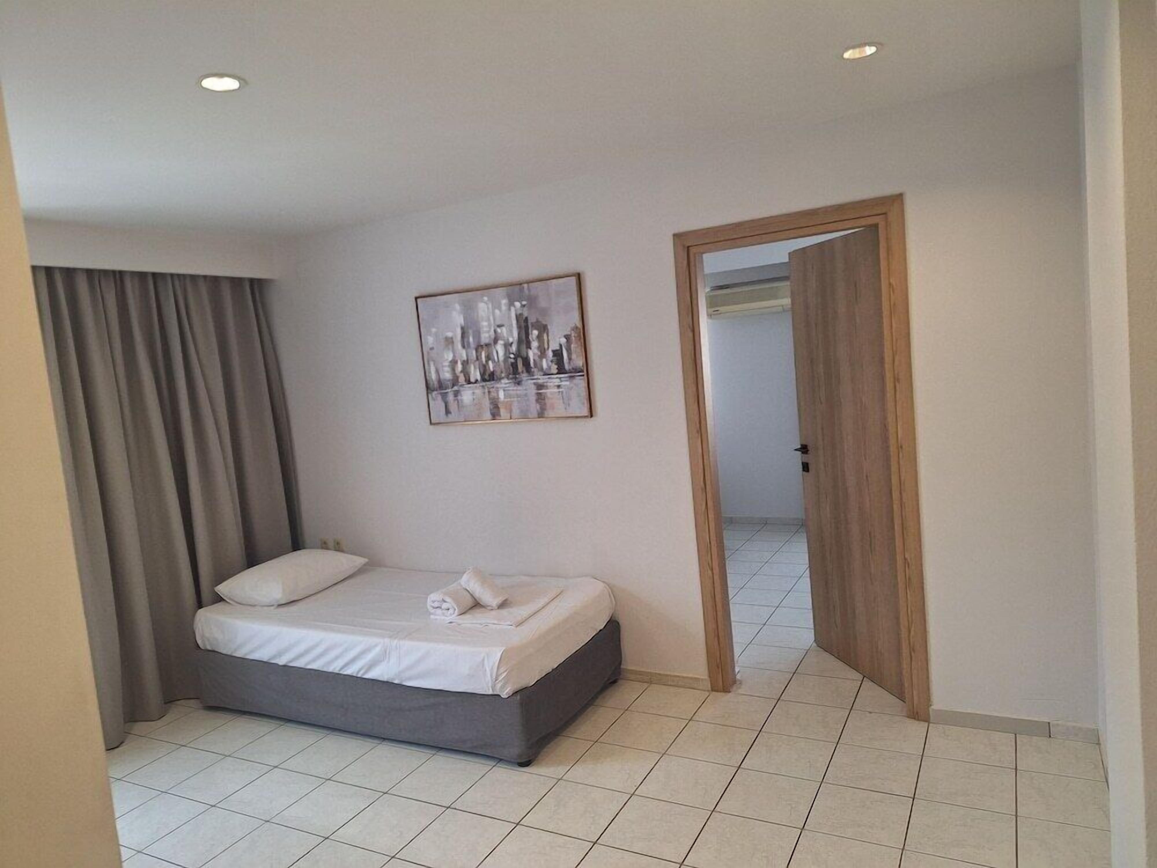 Une chambre d’hôtel avec un lit, un tableau au mur, une porte donnant sur une autre pièce et une fenêtre avec des rideaux.