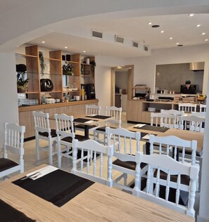 A modern dining area with white chairs and tables, a kitchen in the background, and a person working at a counter. - Soho Blue City Hotel (Hersonissos)