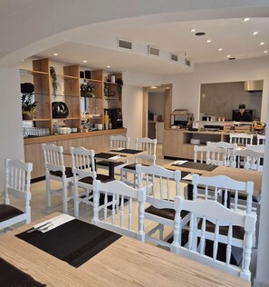 A modern dining area with white wooden chairs and tables, a kitchen area with a person cooking, and shelves with various items. - Soho Blue City Hotel (Hersonissos)