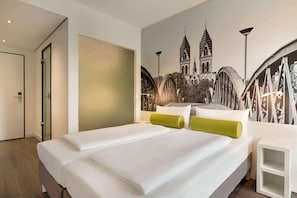 Ett hotellrum med en stor säng, en grön kudde, ett vitt sängbord och en väggmålning som föreställer en bro och en kyrka.